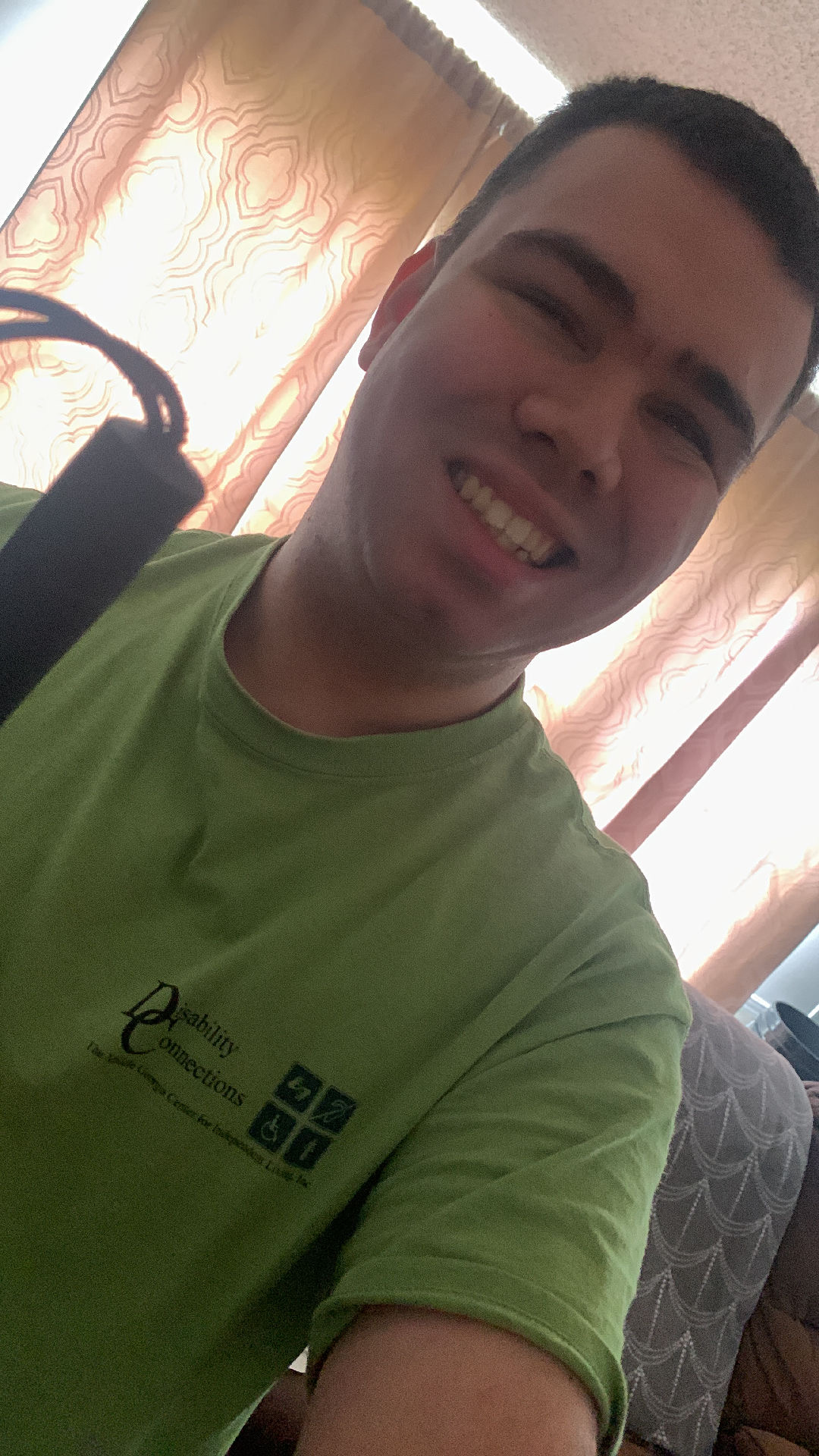 Armando, a young man in the picture, is holding a cane in his right hand, which is partially visible in the foreground. He has a broad smile, short dark hair, and is wearing a lime green t-shirt with a logo on the left side that reads "Disability Connections" with a tagline "The Resource for People with Disabilities" and a graphic of four interlocking squares. In the background, there is a curtain with a floral pattern and a glimpse of a room with a couch. The atmosphere of the picture is warm and cozy, and the man appears to be in a good mood. Copyright 2023 by Armando Vias.