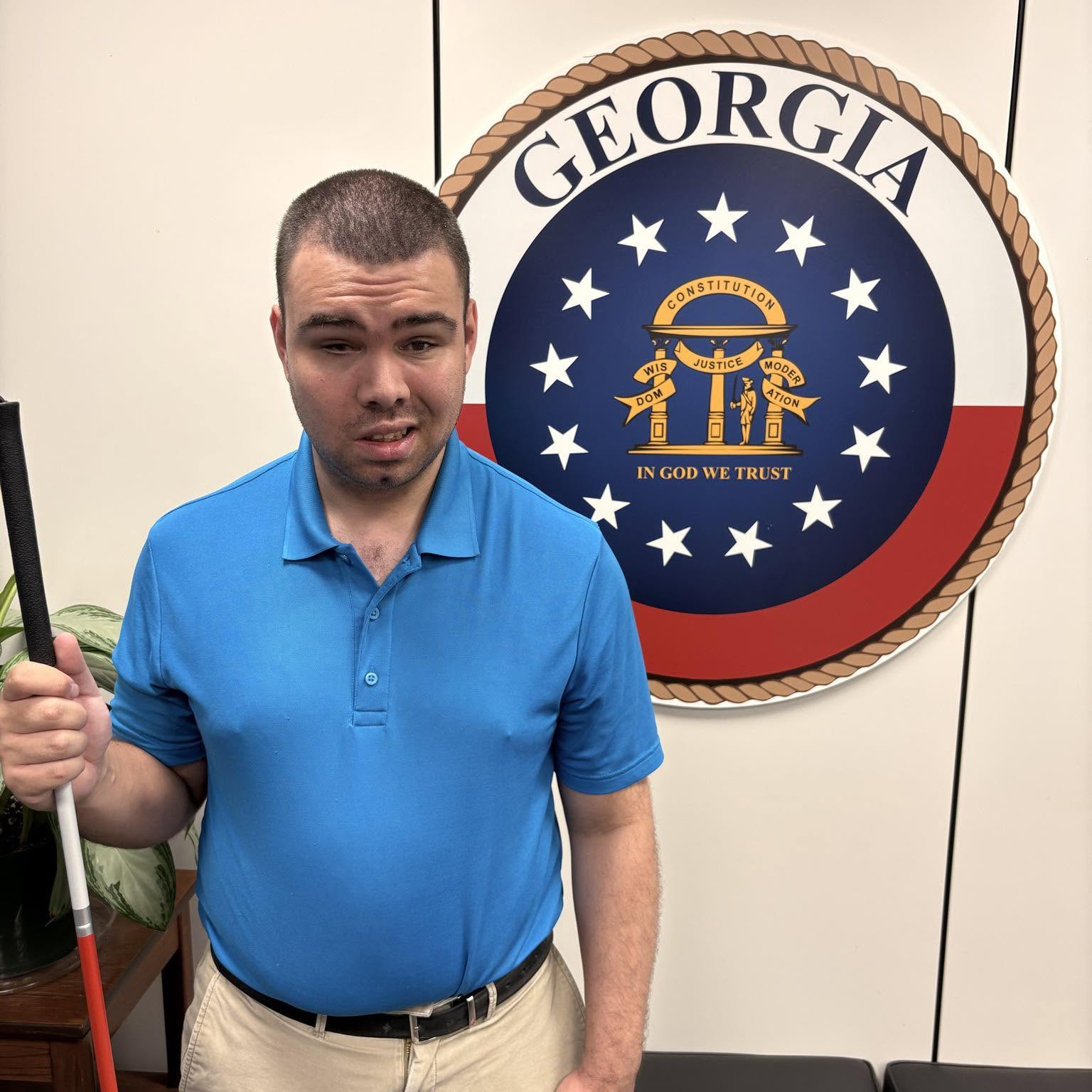 Armando Vias standing outside a Senator's office at the United States Capitol in Washington, D.C. He is wearing a blue polo shirt and khaki pants, holding a white cane in his right hand, with a large Georgia state seal in the background.